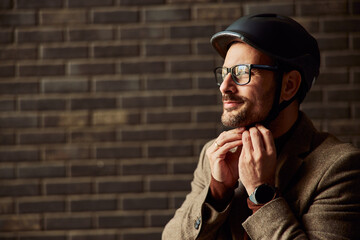 A businessman standing by the brick wall and adjusting a helmet for a bicycle ride.