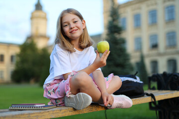 Fototapeta premium Smiling little girl first grader with an apple in her hands at a break on the background of the school.