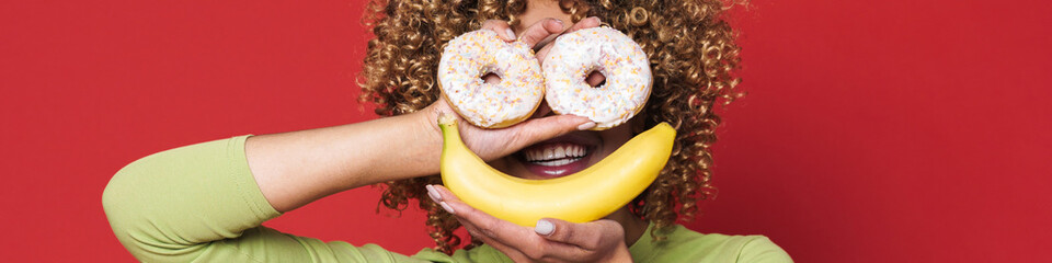Young black woman with afro curls having fun holding donuts and banana