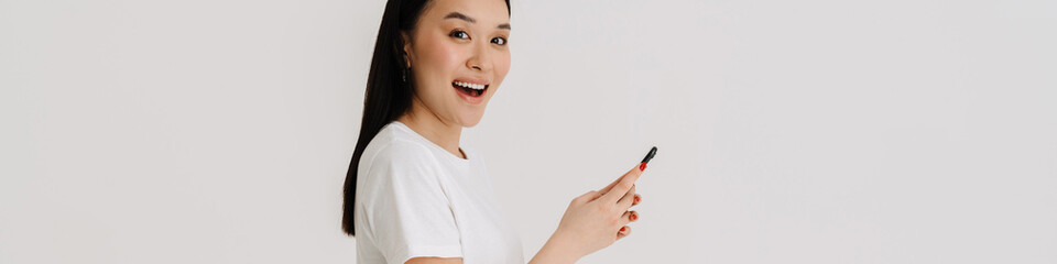 Asian excited woman wearing t-shirt smiling and using cellphone