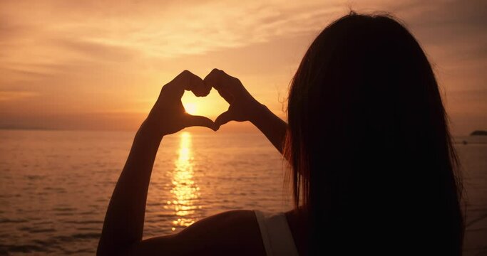 Hands of peaceful woman tourist made a heart shape at sea sunset while traveling at summer vacation. Female hand gesture shows heart symbol silhouette at evening sun. Concept of love to relax at sea.