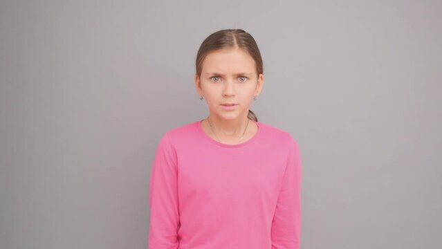 A ten-year-old girl looks at the camera and is angry and shows a feeling of hatred, super slow motion, in the studio on a gray background.