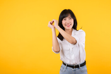 Young female healthcare. Asian woman wearing white shirt feels happy and romantic shapes heart gesture expresses tender feeling poses isolated on yellow background. People affection and care concept.
