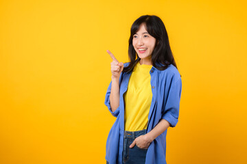 Capture attention with engaging portrait. A young Asian woman wearing a yellow t-shirt and blue shirt showing happy smile while pointing finger to free copy space. education promotion concept.