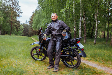 Fototapeta premium Elderly biker leaning on motobike in summer forest. Man in leather clothes relaxing enjoying nature.