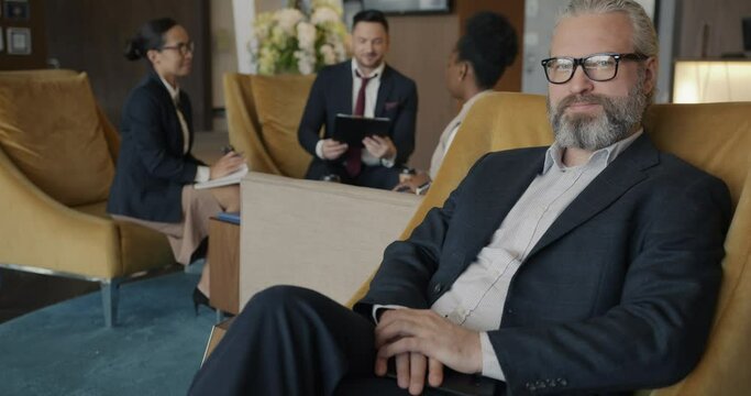 Portrait of successful businessman sitting in hotel lobby and looking at camera while partners diverse group are talking in background. People and occupation concept.
