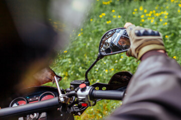 Motorcyclist looking in mirror of motobike outdoors. Man in leather jacket and helmet ride in summer forest.