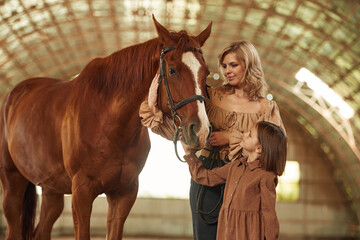 Woman with her little daughter is with horse indoors