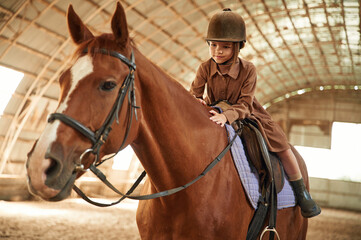 Riding the animal. Cute little girl is with horse indoors