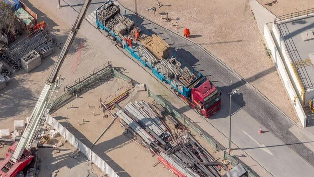 Unloading of building materials by crane at the construction site aerial timelapse. Many workers moving reinforcement. Heavy machinery