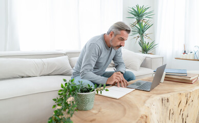 Portrait of Caucasian grey hair man writing notes in notepad while using laptop from workspace in living room at home interior. Work, business and startup planning, brainstorming with device remotely.