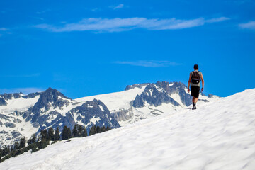 Fototapeta na wymiar Man hiking alone on snow covered trail in summer with blue sky overlooking alpine mountains