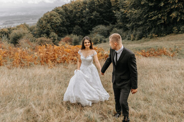 A romantic fairytale couple of newlyweds are walking in a field at sunset, behind fern leaves. The bride is in a white wedding dress, the groom is in a black classic suit and a white shirt