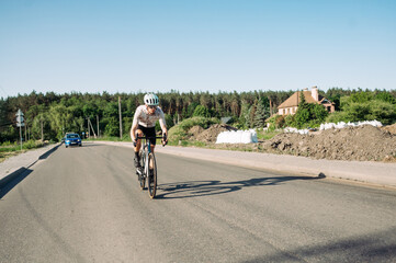 A male cyclist rides a road bike on an asphalt road outside the city on a sunny day, wearing sports equipment.