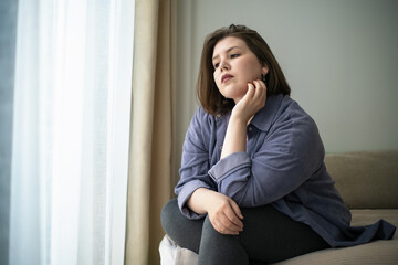 Sad plump or overweight young girl looks out window portrait. Problems of loneliness and mental health plus size people in society. - obrazy, fototapety, plakaty