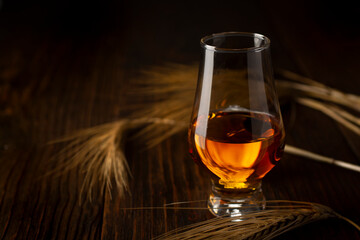 A glass of cognac and a few ears of barley on a dark wooden table.