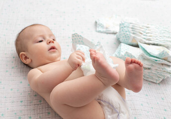 A baby in a diaper lies on a white background plays with a stack of diapers. Copy space for text - obrazy, fototapety, plakaty