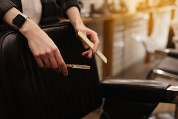 Hair style, hairdresser, beauty and customer service concept. Cropped shot of barber man hands hold professional stylish gold hairbrush and scissors by the workplace, leather black chair in barbershop