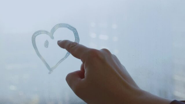Female hand drawing doodle smile face heart on glass wet blue window at home