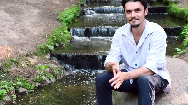 Young, handsome man sits on stone near water in park. Looks at camera and smiles.Man enjoys calmness, murmur of water, unity with nature.Outdoors.Happiness.4k resolution, footage, film, moving image. 