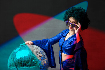 A woman with a disco ball in an afro wig in multi-colored rays of light. Disco style from the...