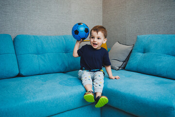 A two-year-old little boy sits on the sofa with a ball in his hands. A developed emotional boy plays with a ball at home