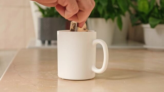 A man mixes sugar with a teaspoon in a white teacup with a tea bag and hot water to make himself tea in the kitchen. Break.