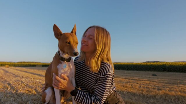 Handheld shot of female teenager feel happy and free with her basenji dog. Young woman kiss a puppy. Golden hour sunset at countryside or farm. Ease of life. Sincere friendship. 