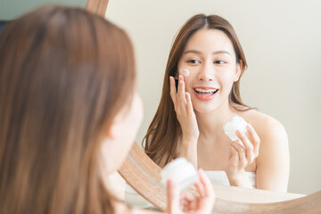 Facial beauty skin care, smile of pretty asian young woman in bathrobe looking at mirror, hand applying moisturizer lotion on her face, holding jar of skin cream before makeup cosmetic routine at home
