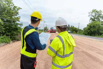 Asian engineer holding tablet and foreman holding road construction blueprints, on construction site to supervise road construction In the background is a road under construction and machinery working