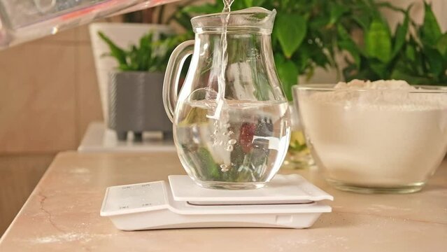 A woman measures water into a glass jug standing on an electronic scale. Preparation of yeast dough for pizza.