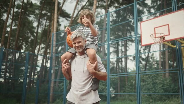 Сute little boy is sitting on dad's shoulders and holding basketball. Loving father and son smiling, posing and looking at camera on background of basketball court. Family spending time together.