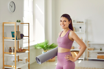 Fototapeta na wymiar Happy slim smiling woman in sportswear going to do sport exercises, holding yoga mat and looking cheerful at camera in the living room at home. Workout sport and body care concept.