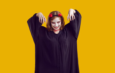 Stale corpse rises from the grave to participate in Halloween party. Woman in black robe, makeup, devil horns, bloody trail on her neck scares audience with her hands up. Isolated on yellow.