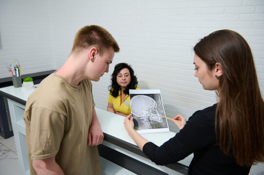 Young woman dentist doctor explaining X-ray image to a male patient, standing together at the reception counter of a modern medical clinic or outpatient hospital. Healthcare and medicine. Diagnostics