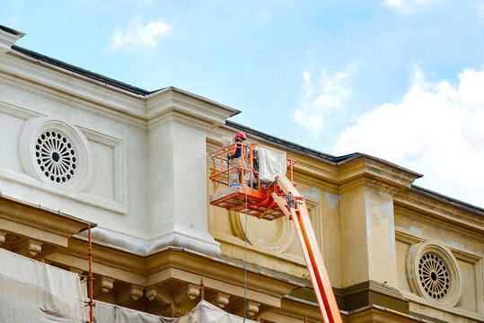 Worker on aerial platform restore, repair historic facade of old building. Man in cradle, restoring plaster on facade. Worker painting building, external building work. Man paint building wall