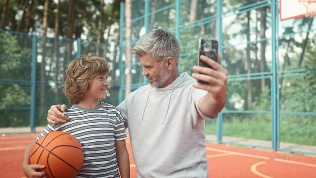 Smiling father with little son taking selfie picture with smartphone on background of basketball court. Cute teenage boy holds basketball and posing for selfi with his dad. Happy childhood concept.