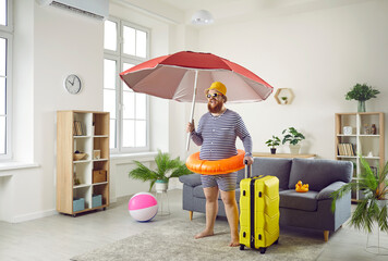 Funny excited chubby man standing at home is finally ready to go on long-awaited summer vacation. Joyful man with suitcase, inflatable circle and large beach umbrella stands in middle of living room. - obrazy, fototapety, plakaty