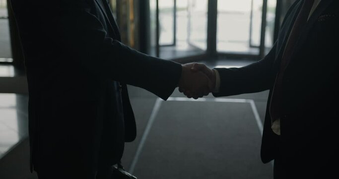 Close-up of male entrepreneurs shaking hands and gesturing in modern glass wall hotel. Businessmen are discussing agreement together.