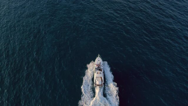 Tilt shot from top on luxurious white yacht cruising in blue ocean waters in sunset. Aerial drone shot of group of friends have party on deck of fancy sail boat in sea