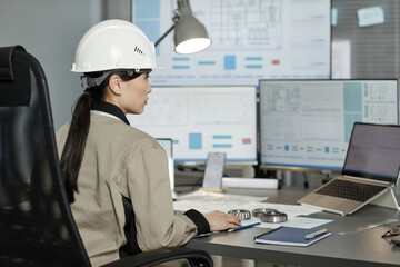 Side view portrait of female engineer wearing hardhat at workplace in office and using computer...