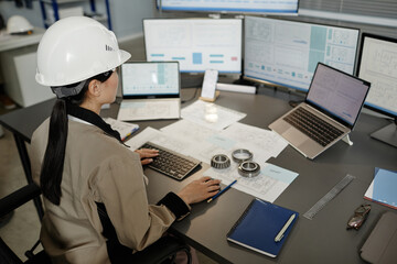 High angle portrait of female engineer wearing hardhat at workplace in office and using multiple...