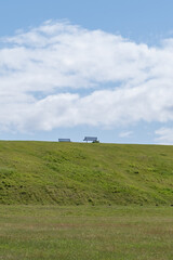 Fototapeta premium Two public bench chairs sitting on top of a grass embankment on a bright cloudy day with some blue Skys