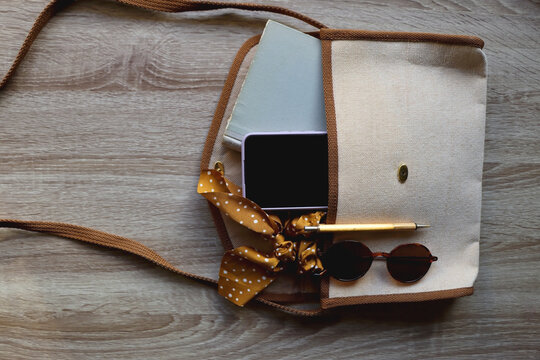 Neutral purse with vintage book, mobile phone, golden pen, polka dot scrunchie and round sunglasses. Top view, wooden background.