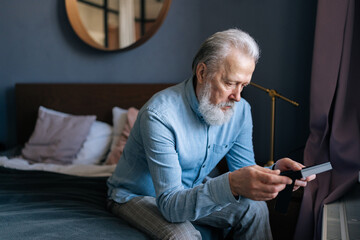 Medium shot of unhappy bearded mature male looking at frame of photo, stroking picture with sadness from missing someone, sitting on bed. Concept of nostalgia, grief, longing and loneliness in old age