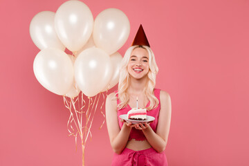 Happy european lady in party hat posing near balloons and holding piece of cake with candle, making...