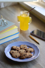 Plate of chocolate chip cookies, stack of books, reading glasses, orange soda, mobile phone and pen on the table. Hygge at home. Pastel colors, selective focus.
