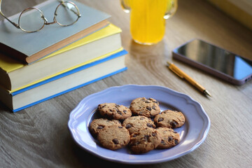 Plate of chocolate chip cookies, stack of books, reading glasses, orange soda, mobile phone and pen on the table. Hygge at home. Pastel colors, selective focus.