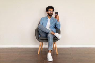 Great App. Young Indian Man Using Smartphone While Sitting In Chair Indoors