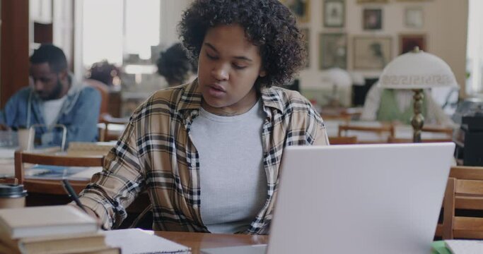Confident African American woman student working with laptop searching information and writing in university library. Modern technology and education concept.